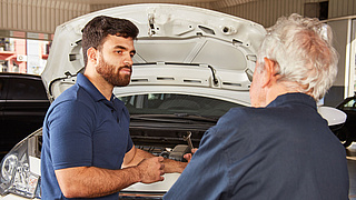 Ein jüngerer und ein älterer Automechaniker vor einer geöffneten Motorhaube.