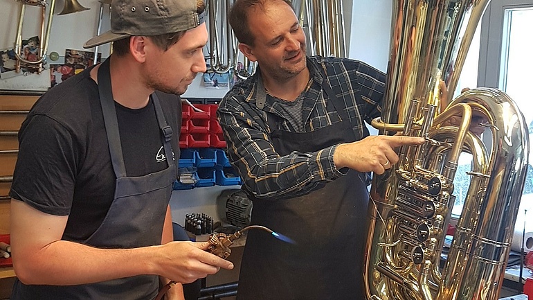Ein Metallblasinstrumentenmachermeister zeigt einem Meisterschüler in seiner Werkstatt etwas an einem Metallblasinstrument.