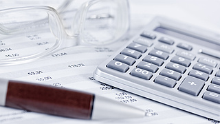 Brille, Kugelschreiber und Taschenrechner liegen auf einem Papier mit Zahlen.