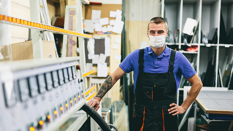 Ein Handwerker mit Mundschutz in einer Glaserei blickt in die Kamera. Im Hintergrund sind in Fächer sortierte zugeschnittene Glasstücke zu sehen.