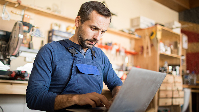 Handwerker sieht in einer Werkstatt konzentriert etwas auf einem Laptop an.