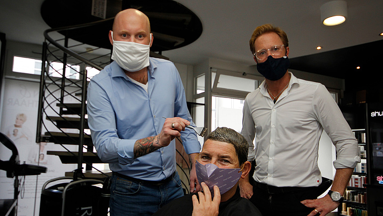 Die beiden Friseure Daniel und Marco Illiges stehen hinter einer Kundin in ihrem Laden, alle tragen Gesichtsmasken.