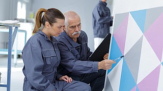 Ein älterer Maler mit einer jüngeren Malerin in der Hocke vor einer mit farbigen Dreiecken bemalten Fläche.