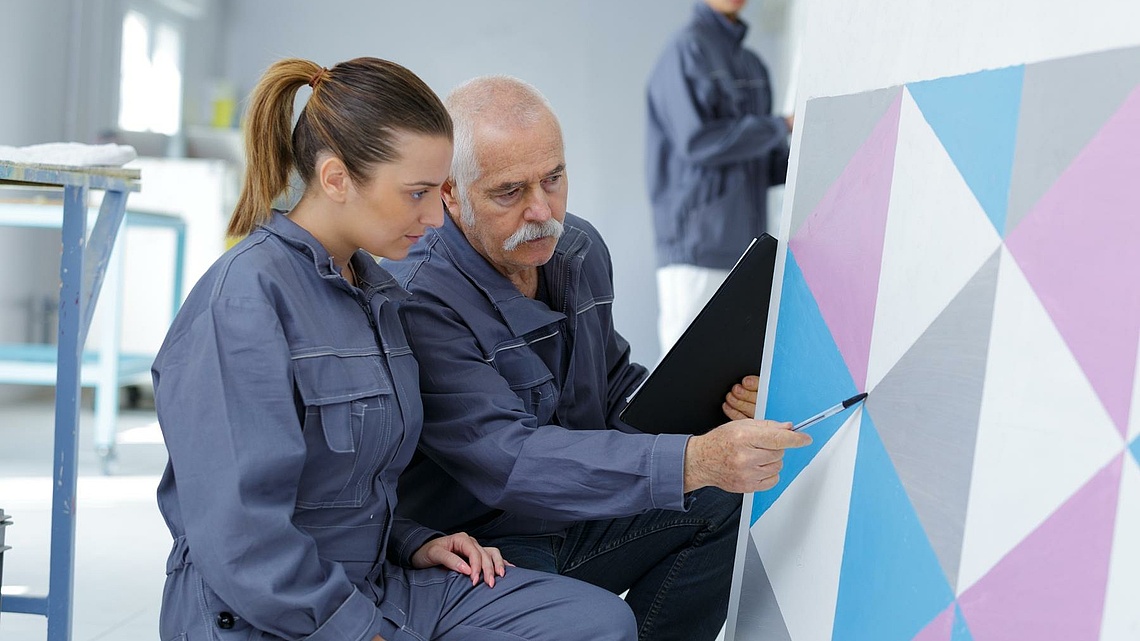 Ein älterer Maler mit einer jüngeren Malerin in der Hocke vor einer mit farbigen Dreiecken bemalten Fläche.