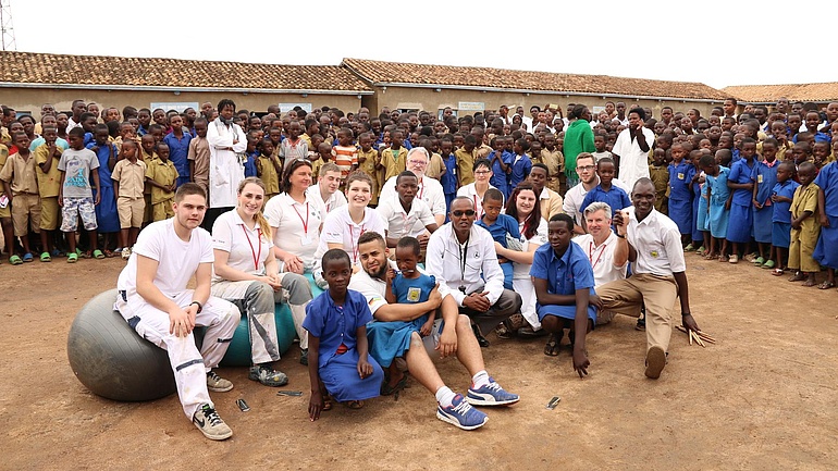 Deutsche Handwerker im Hof einer afrikanischen Schule sind umringt von afrikanischen Schulkindern in blauen Schuluniformen.