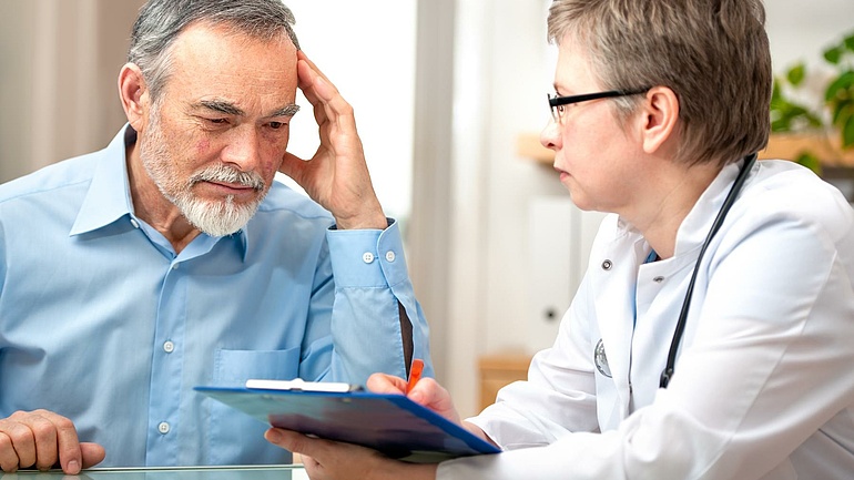 Älterer Herr im hellbauen Hemd im Gespräch mit einer Ärztin, die ein Tablet in den Händen hält.