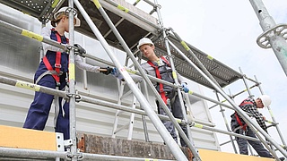 Drei Gerüstbauer auf einem Gerüst mit Sicherheitsausrüstung.