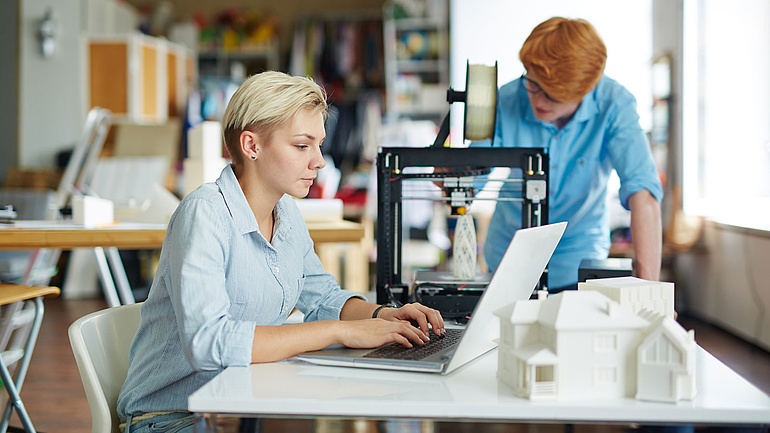 Zwei junge Menschen fertigen Architekturmodelle an einem 3-D-Drucker.