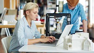 Zwei junge Menschen fertigen Architekturmodelle an einem 3-D-Drucker.