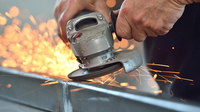 Zwei Hände schleifen ein Metallstück mit einer Schleifmaschine. Dabei sprühen Funken.