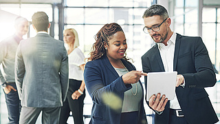 Aufnahme von zwei Kollegen, die sich über ein digitales Tablet in einem Büro unterhalten.