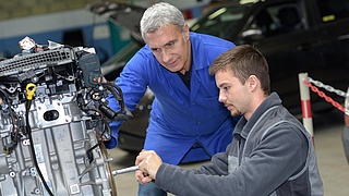 Ein Azubi repariert etwas an einer großen Maschine, ein Ausbilder schaut zu.