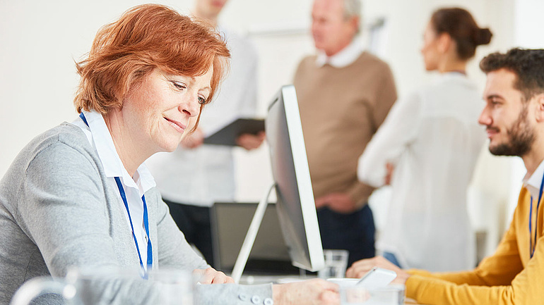 Eine Frau am Smartphone und ein Mann am Monitor an ihren Büroarbeitsplätzen, im Hintergrund unterhält sich eine Gruppe von Menschen.
