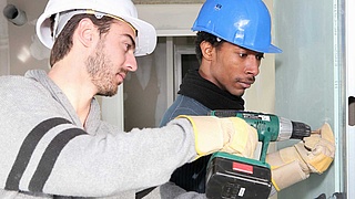 Zwei Handwerker mit Schutzhelmen befestigen eine Platte an einer Wand.