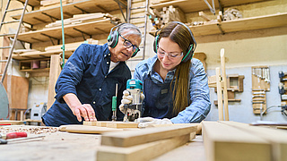 Junge Frau an der Stichsäge, älterer Handwerker schaut ihr dabei über die Schulter