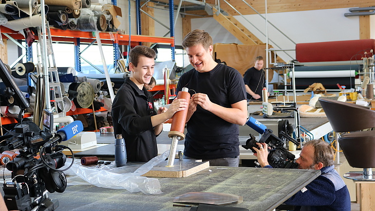 Zwei junge Männer bauen etwas in einer Werkstatt, ein Kameramann filmt sie dabei.