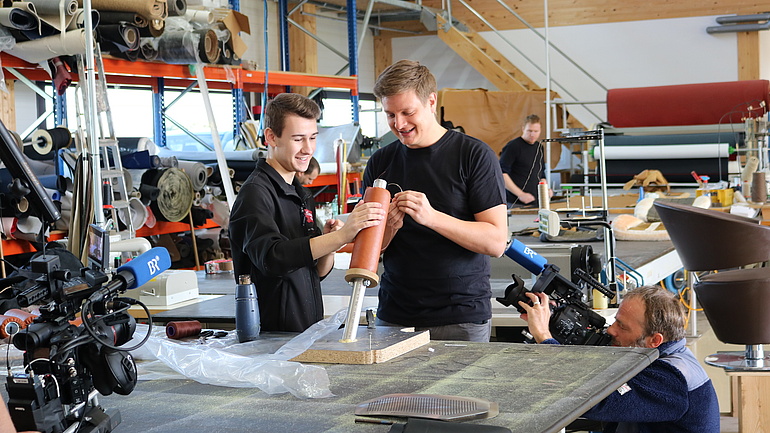 Zwei junge Männer bauen etwas in einer Werkstatt, ein Kameramann filmt sie dabei.