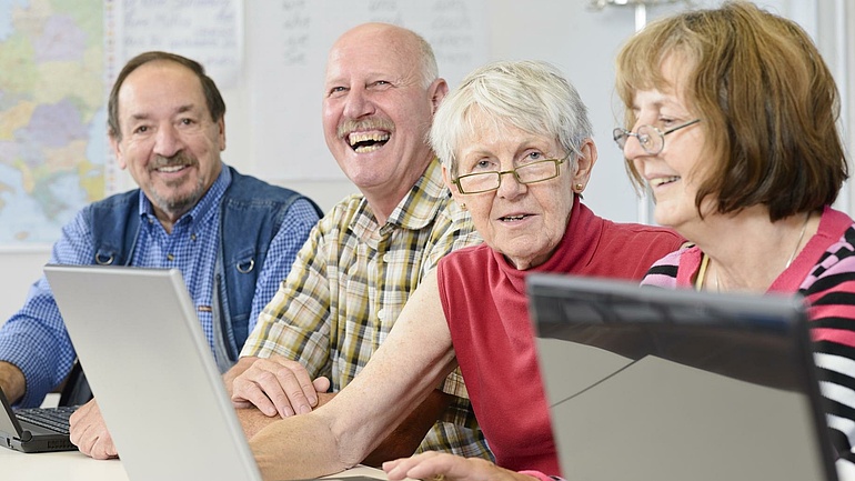Lachende Senioren sitzen nebeneinander an einem Tisch und arbeiten mit Laptops.