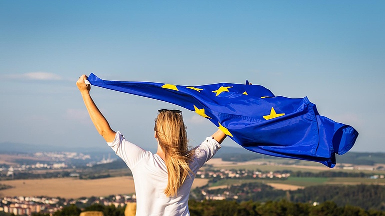 Eine junge Frau steht auf einem Hügel vor einem Kornfeld und hält eine Europaflagge in den Wind.