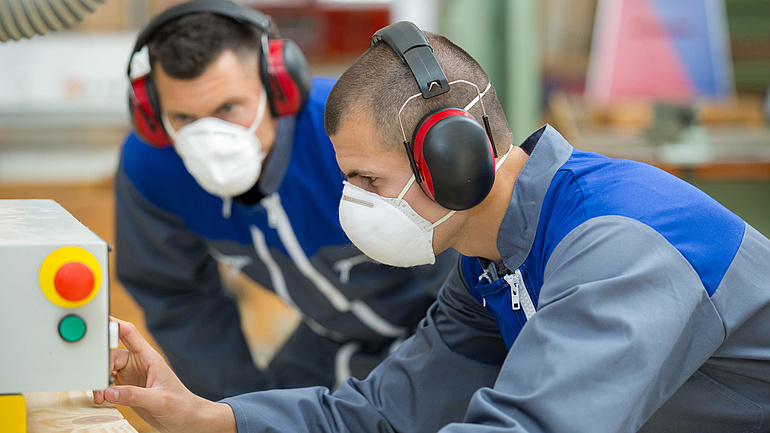 Zwei Handwerker in Blaumännern mit Gehör- und Mundschutz überprüfen konzentriert eine elektronische Anzeige.