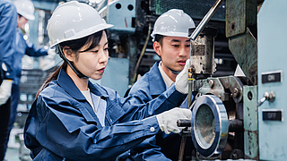 Zwei junge asiatische ArbeiterInnen in blauer Schutzkleidung mit Helm bedienen eine Maschine in einer Werkhalle.