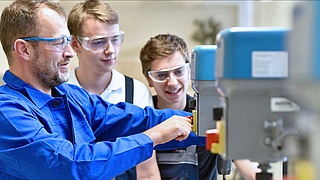Handwerker mit zwei Auszubildenden bei der Unterweisung an einer Maschine.
