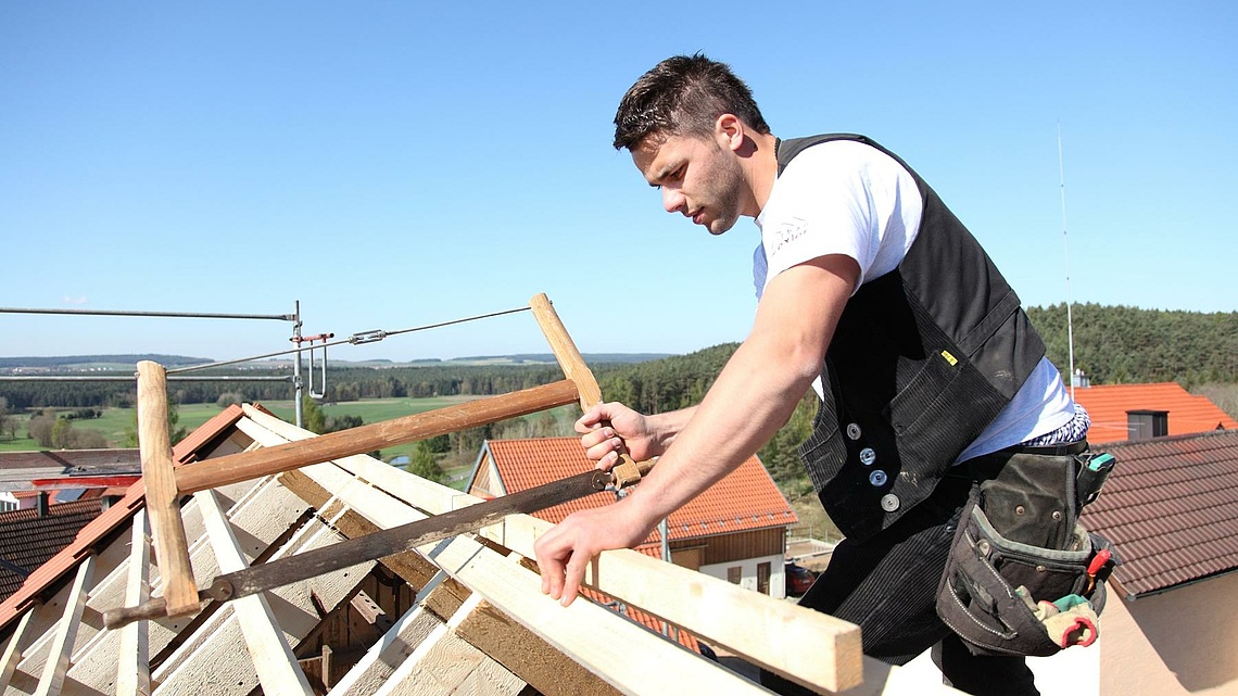 Ein Zimmermann sägt ein Stück Holz beim Ausbau eines Daches, blauer Himmel im Hintergrund.