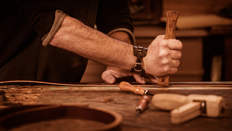 Bildausschnitt der Hand eines Sattlers, der einen Lederriemen mit einem Holzstück bearbeitet.