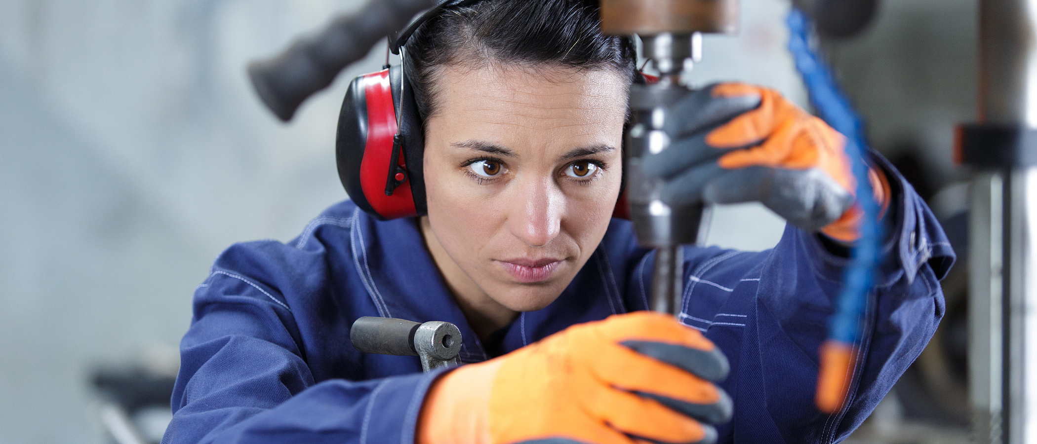 Handwerkerin mit roten Ohrenschützern und orangen Handschuhen konzentriert sich auf ihre Arbeit an einer Drechselmaschine.