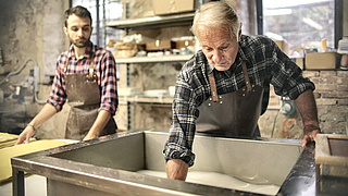 Ein älterer und ein jüngerer Handwerker, beide tragen Karohemden, arbeiten an einer Metallwanne mit einer weißen Flüssigkeit.