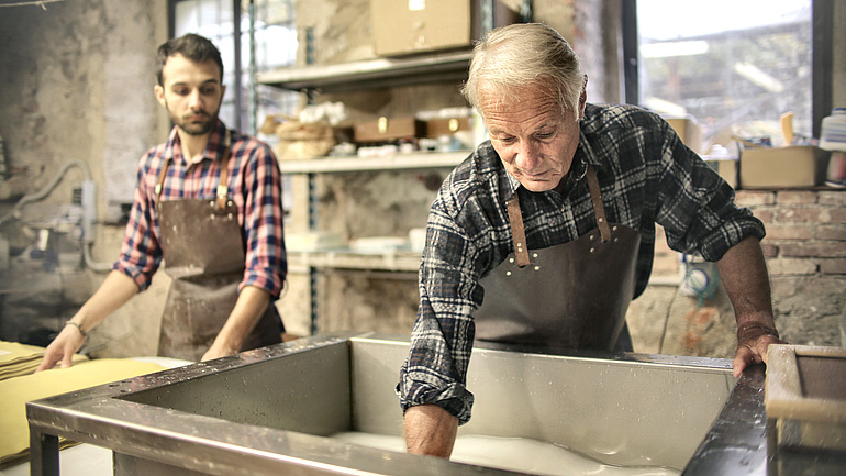 Ein älterer und ein jüngerer Handwerker, beide tragen Karohemden, arbeiten an einer Metallwanne mit einer weißen Flüssigkeit.