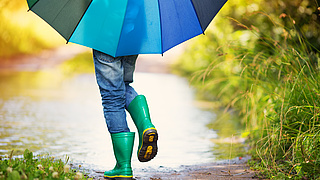 Ausschnitt eines Kindes mit Gummistiefeln und einem bunten Regenschirm.