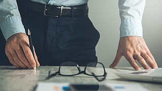 Mann steht an seinem Arbeitsplatz mit einem Stift in der Hand, vor ihm Zettel, Laptop und eine Brille.