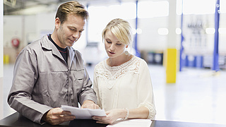 Ein Mechniker und eine junge Frau stehen an einem Tresen und betrachten gemeinsam ein Stück Papier.