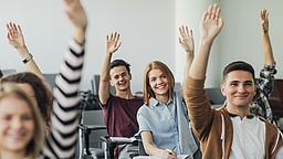 Junge Menschen in einer Schulklasse heben die Hand.