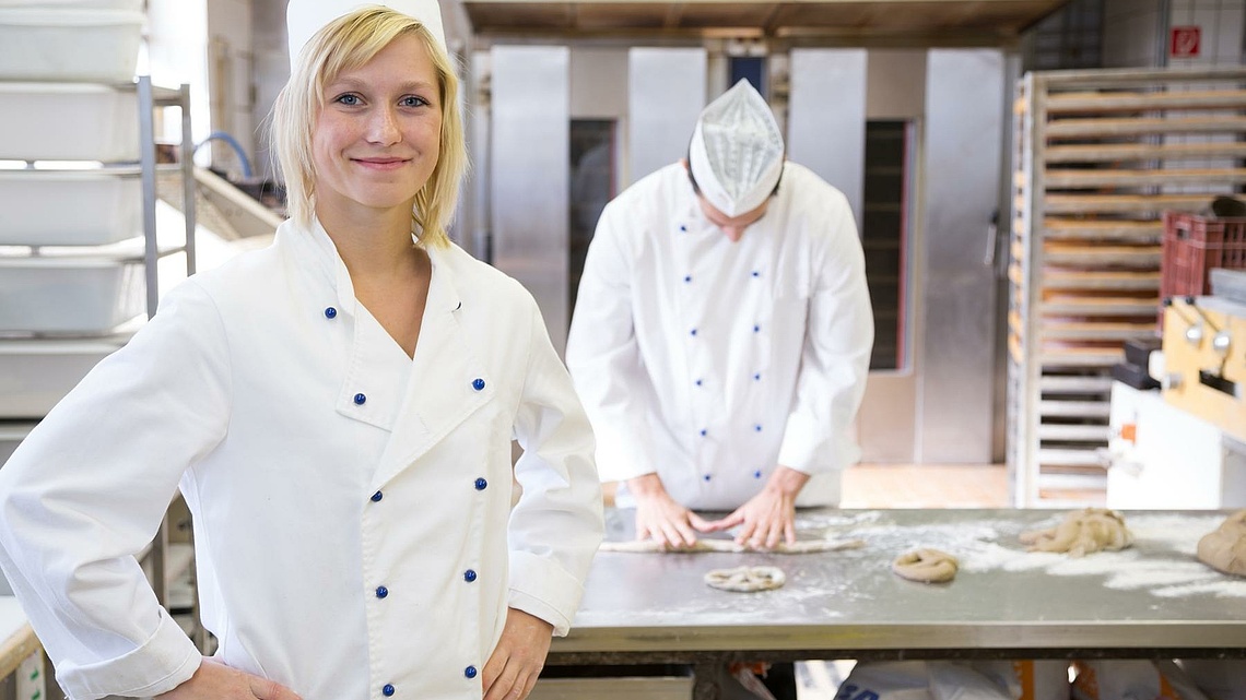 Eine blonde Bäckerin präsentiert sich in ihrer Backstube. Im Hintergrund arbeitet ein Bäcker an einem Metalltisch.