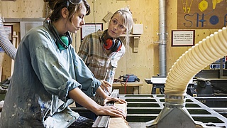 Ausbilderin und Auszubildende stehen an einer Sägemaschine in einer Tischlerei.