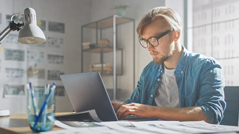 Junger Mann mit Brille sitzt zu Hause mit Laptop an einem Schreibtisch.