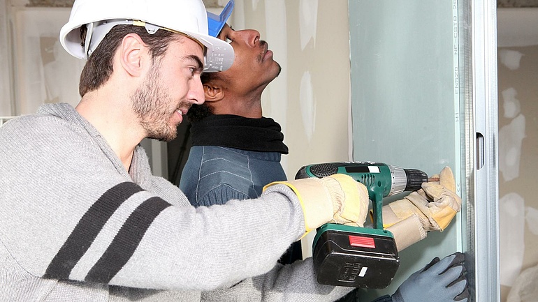 Zwei junge Handwerker mit Bauhelmen montieren eine Wandplatte mit einem Akkuschrauber.