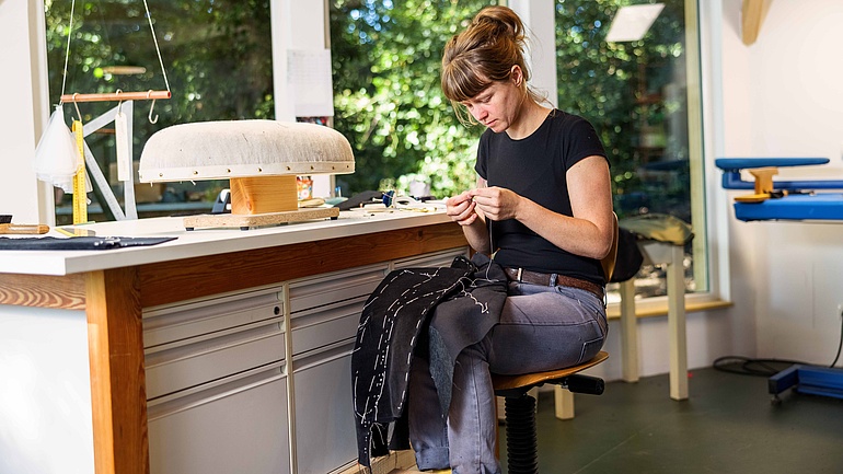 Die Schneiderin Anna Meyer sitzt in ihrem Atelier vor ihrem Werktisch und prüft eine Näharbeit.