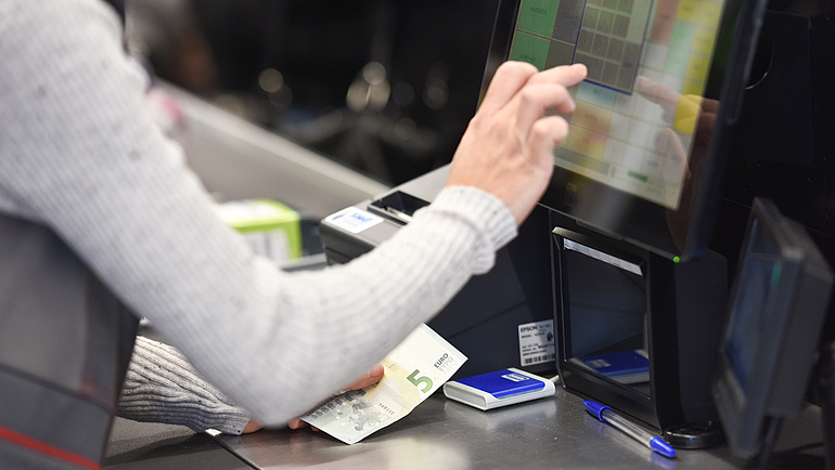 Ausschnitt einer Kasse, vor der eine Person mit einem Geldschein in der Hand steht und etwas eintippt.