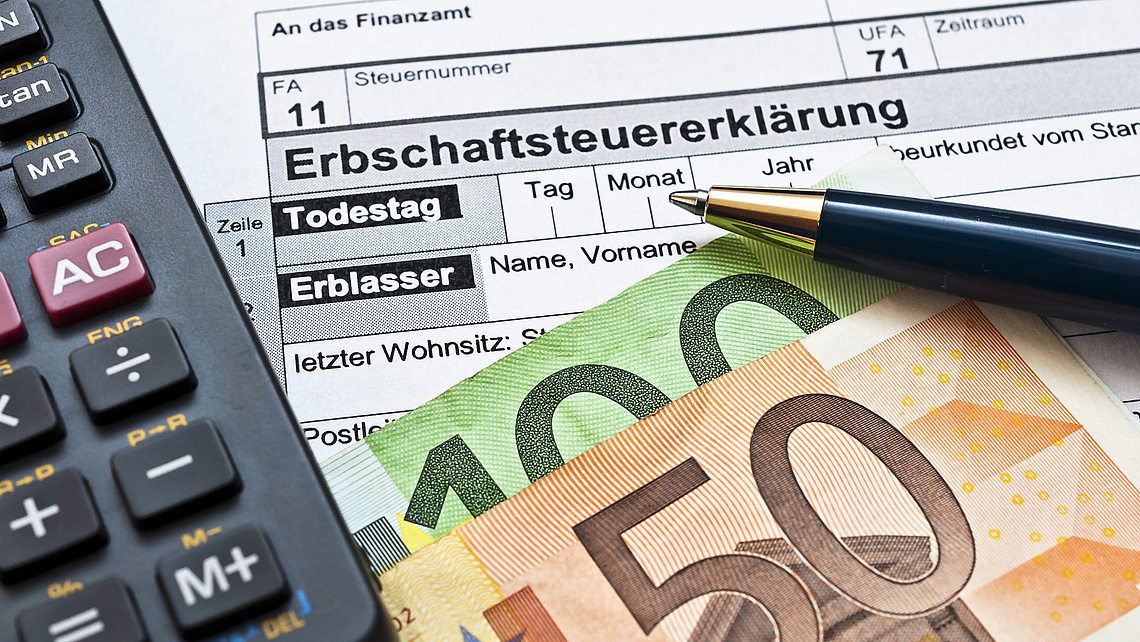 Neben einem Taschenrechner liegen Geldscheine und ein Kugelschreiber auf einem Erbschaftsteuererklärungsformular.