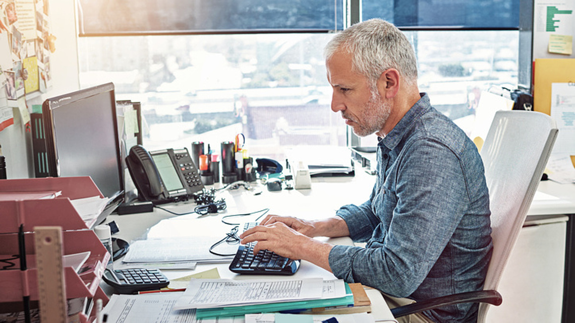 Ein Mann sitzt an einem Büroschreibtisch mit zahlreichen Unterlagen, Telefonanlage, Tatstatur und Monitor.