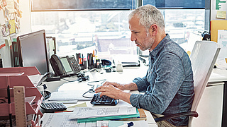 Ein Mann sitzt an einem Büroschreibtisch mit zahlreichen Unterlagen, Telefonanlage, Tatstatur und Monitor.