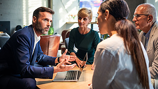 Besprechung an einem Tisch: Ein Mann im blauen Anzug gestikuliert und wendet sich einer kleinen Gruppe von Menschen zu.