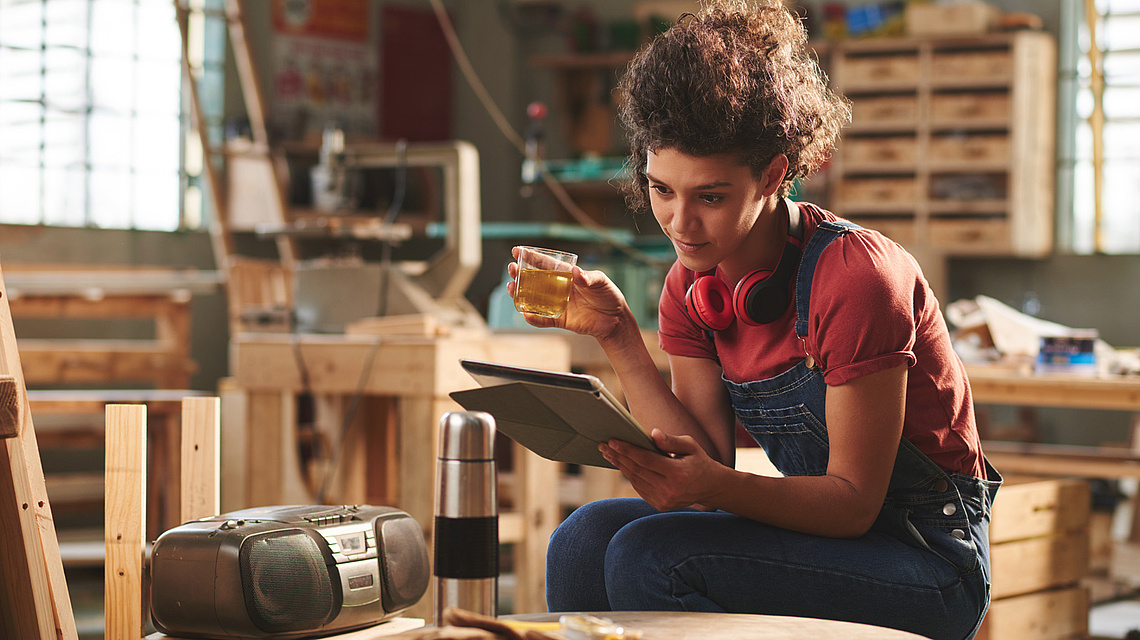 Eine junge Frau sitzt mit einem Tablet in einer Werkstatt, vor ihr auf dem Tisch steht eine Musikanlage.