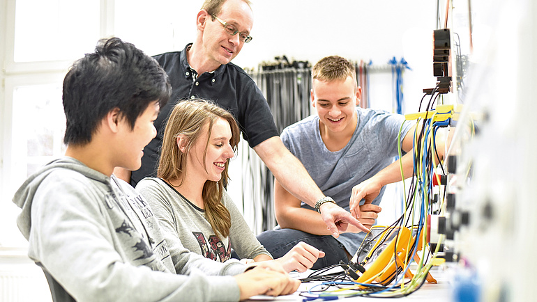 Drei junge Menschen betrachten ein Messgerät an das verschiedenfarbige Kabel angeschlossen sind. Hinter ihn steht ein Ausbilder, der den Apparat erklärt.
