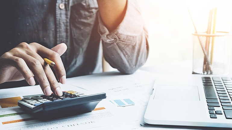 Ausschnitt eines Mannes am Schreibtisch, der auf einem Taschenrechner tippt, ein Laptop daneben.