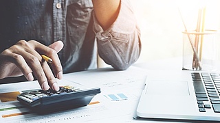 Ausschnitt eines Mannes am Schreibtisch, der auf einem Taschenrechner tippt, ein Laptop daneben.