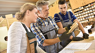 Ein Mesiter erklärt seinen Gesellen etwas am Beispiel zweier Holzscheite.